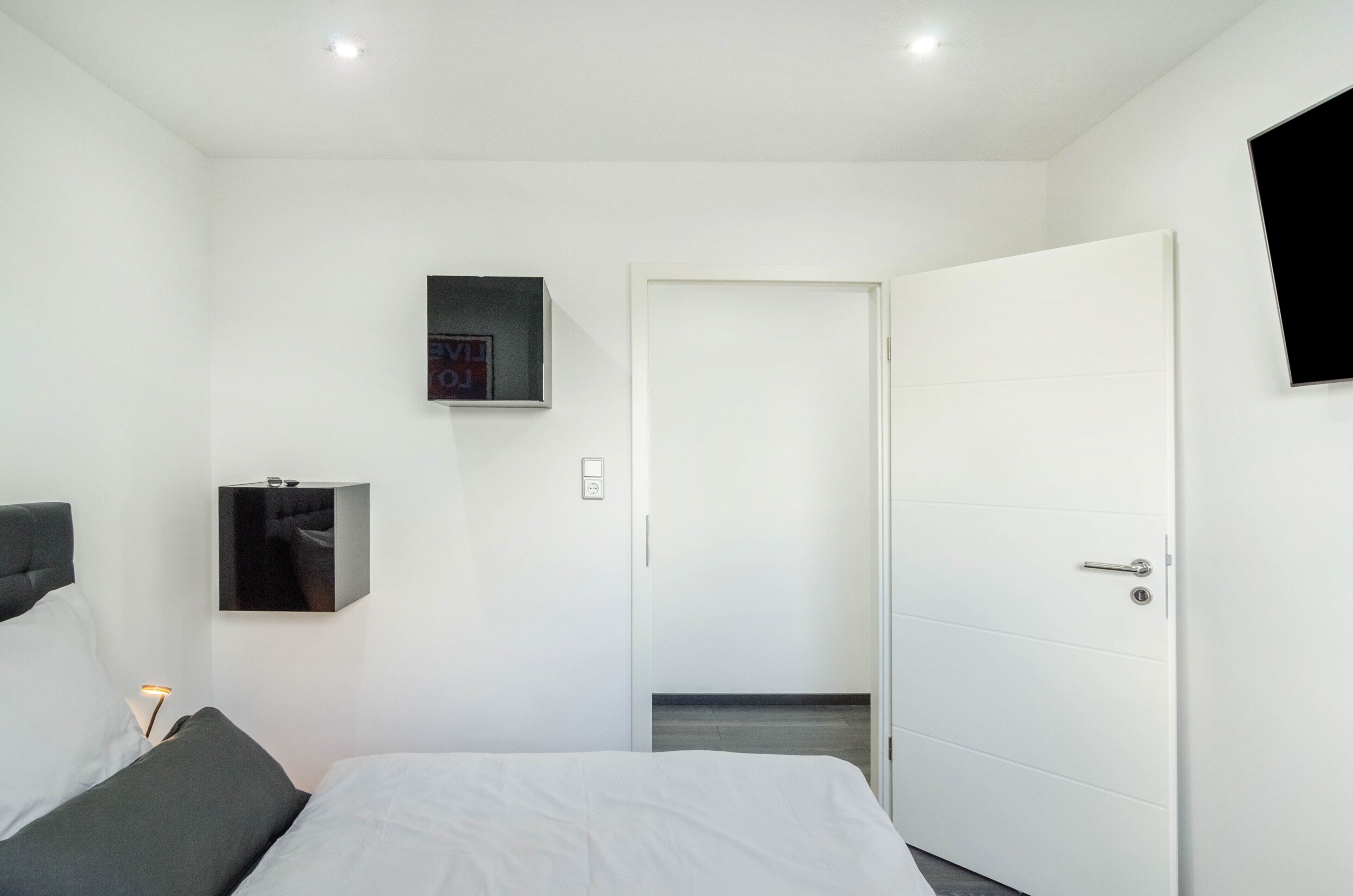 Ein modernes Hotelzimmer mit Bett, Nachttisch mit Lampe, Kleiderschrank und einer Tür zu einem weiteren Raum.