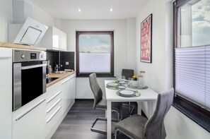 Une cuisine compacte dotée d’appareils électroménagers modernes, un coin repas avec une table pour deux personnes et une fenêtre avec des stores.
