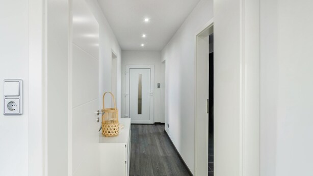 Un couloir aux murs blancs, avec un plancher en bois, un panier posé sur un comptoir et une porte au fond.