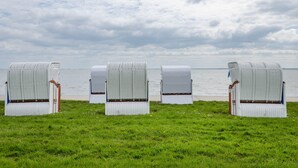 Quatre chaises en osier blanc, dotées d’une structure en bois et d’assises marron, sont disposées sur une pelouse, offrant une vue sur la mer et un ciel nuageux.