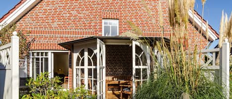 A brick house with a white-framed window and door, a wooden bench outside, and tall grasses in the foreground.