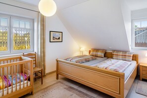 Ein Schlafzimmer mit einem hölzernen Bett, einem Reisebettchen, einem Nachttisch, einer Lampe und einem Fenster mit Blick auf Bäume.