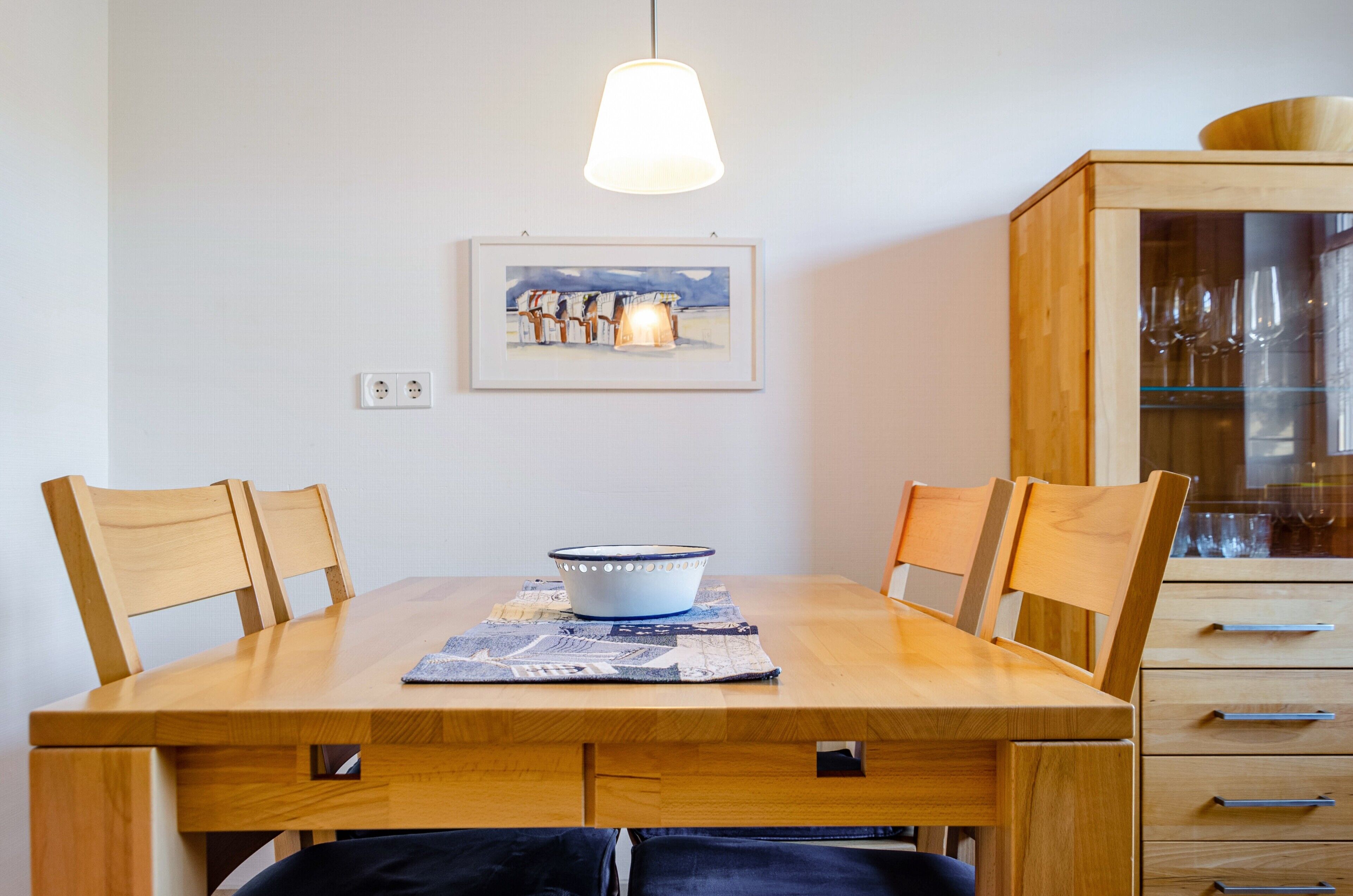 A dining table with chairs, a bowl, a placemat, a framed picture, and a wooden cabinet with glassware.