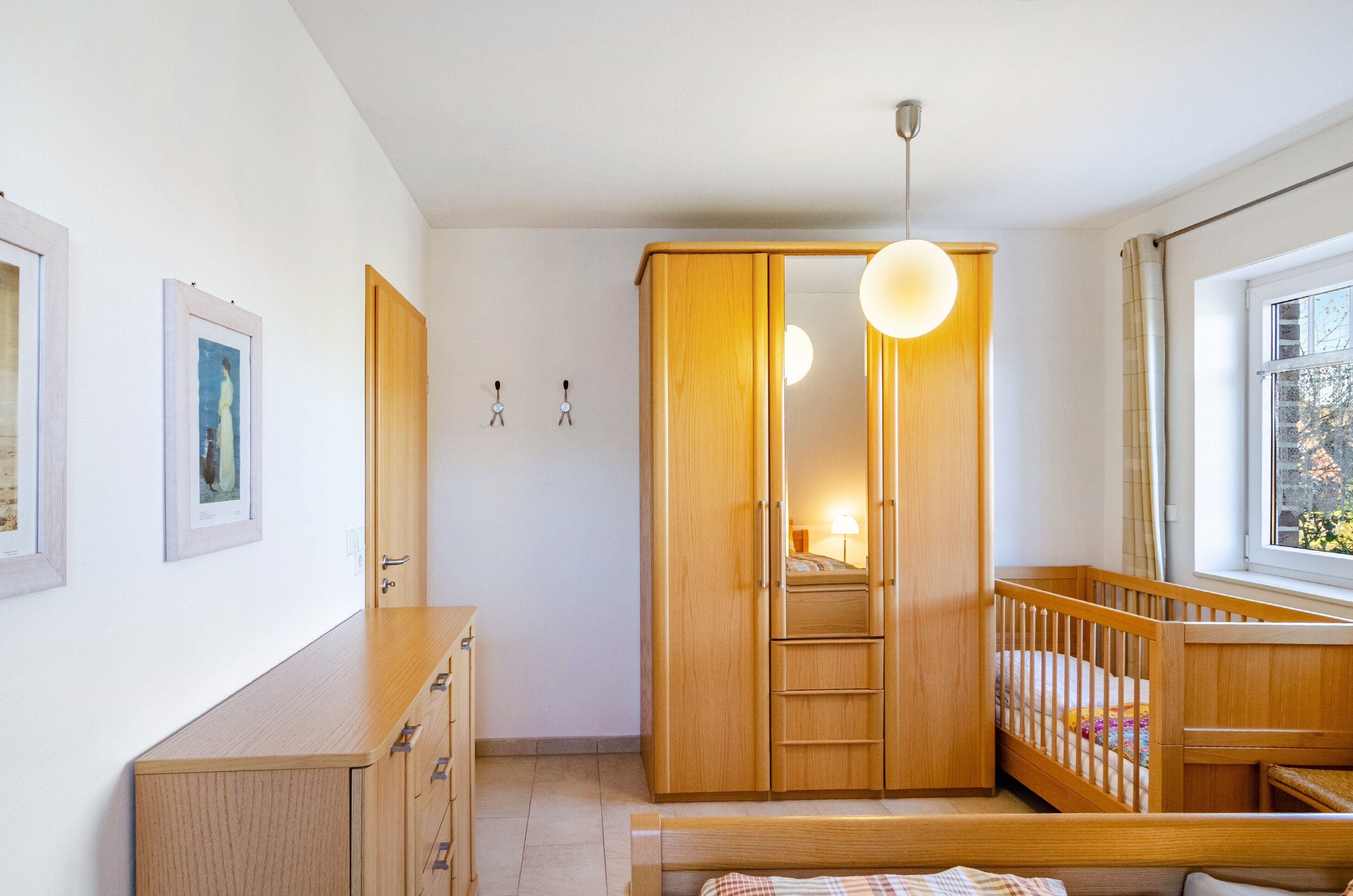 A bedroom with a wooden wardrobe, a crib, a bedside table, and a window with a view.