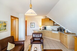 Un salon avec un canapé en cuir marron, une table basse en verre et un tapis marron. On aperçoit un coin repas avec une table et des chaises en bois. La cuisine est équipée de meubles en bois et d’un four à micro-ondes.