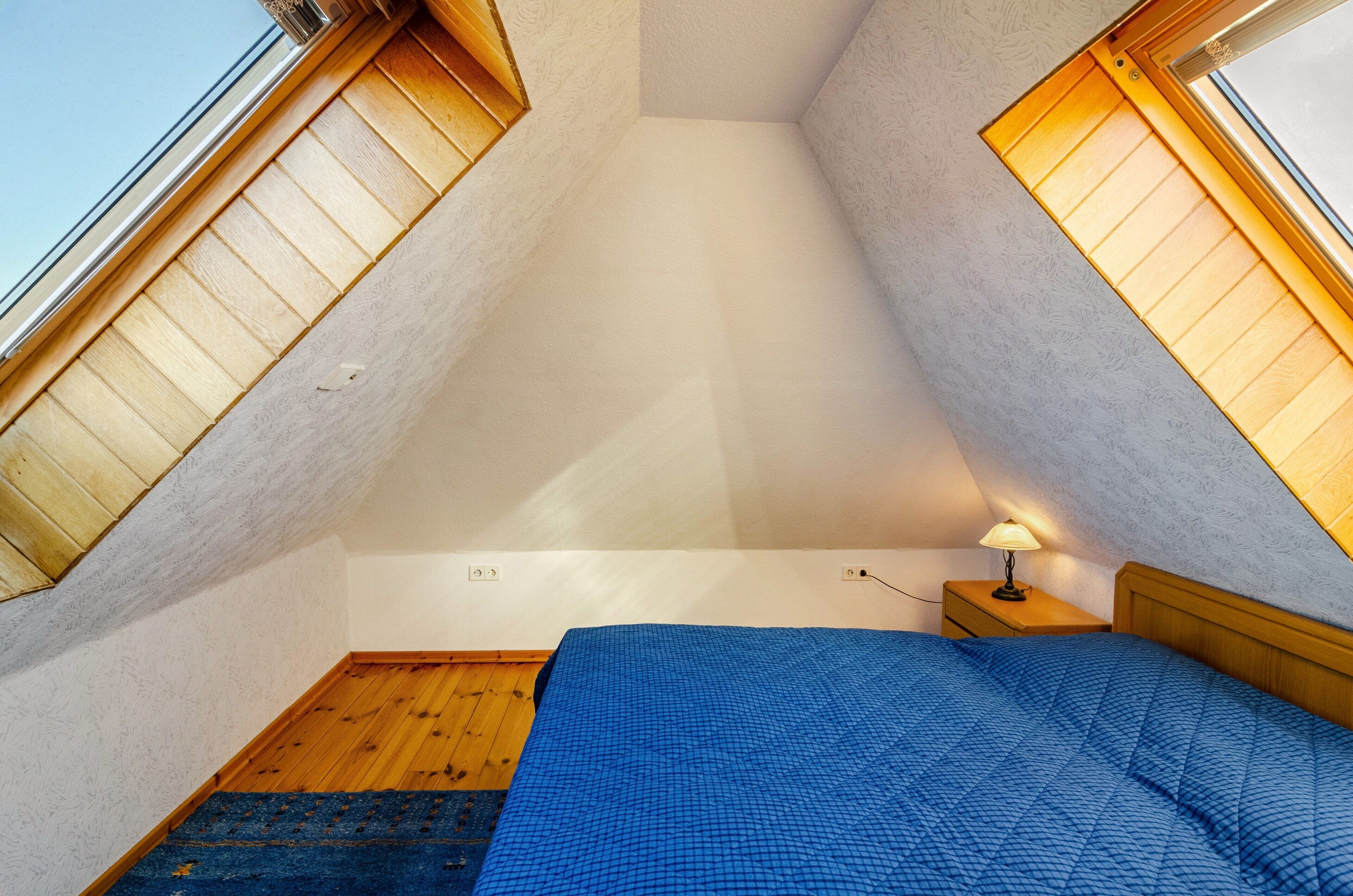 Une pièce avec un plafond en pente, des murs en bois, un lit recouvert d’une couverture bleue, une table de chevet avec une lampe et une fenêtre dotée d’un store en bois.