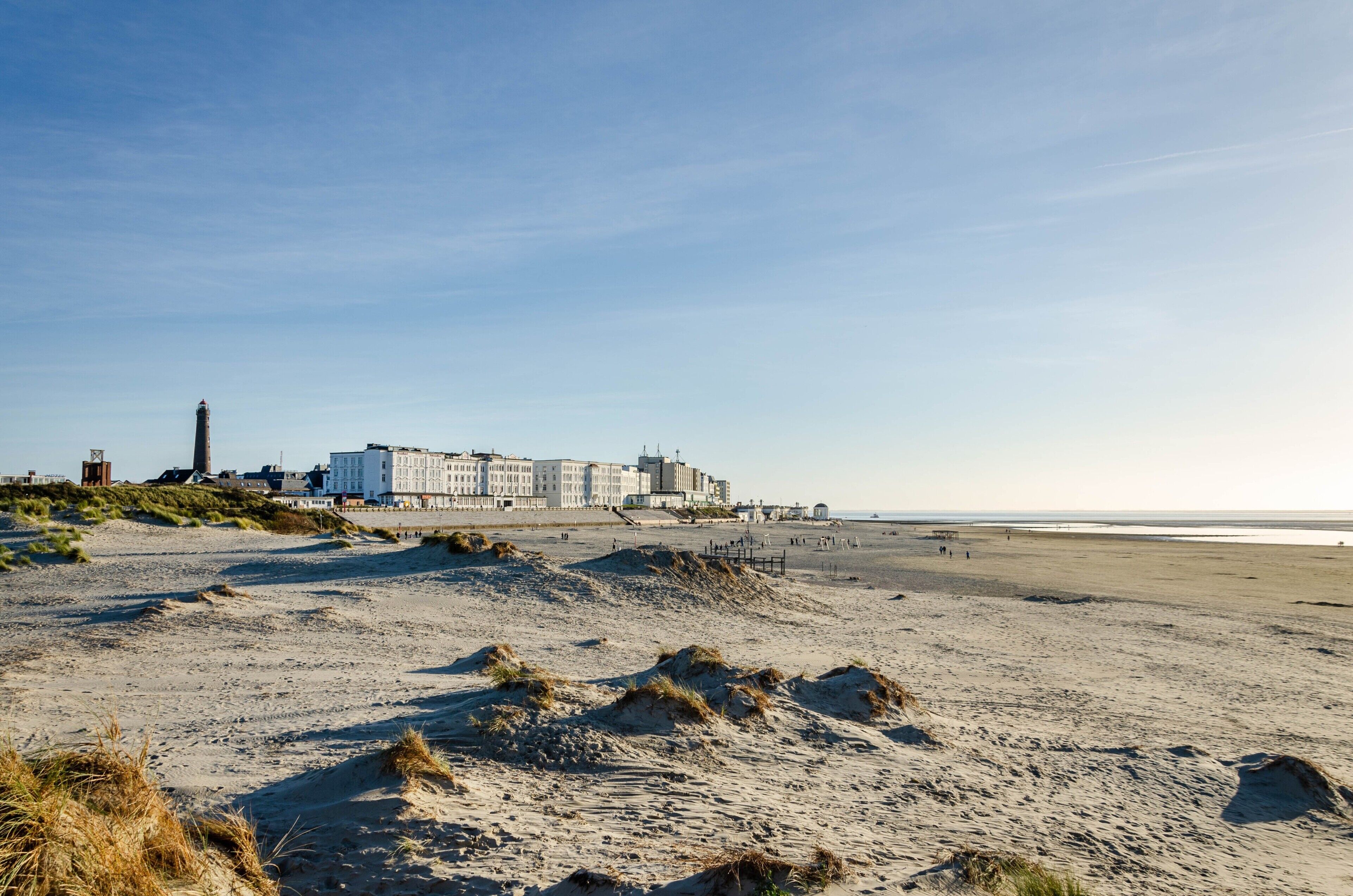 Un hotel frente al mar con vista a la playa, dunas de arena y un faro al fondo.
