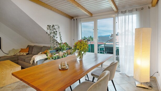 Zona pranzo con tavolo in legno, sedie, un vaso con fiori, un divano e un'ampia finestra con tende.