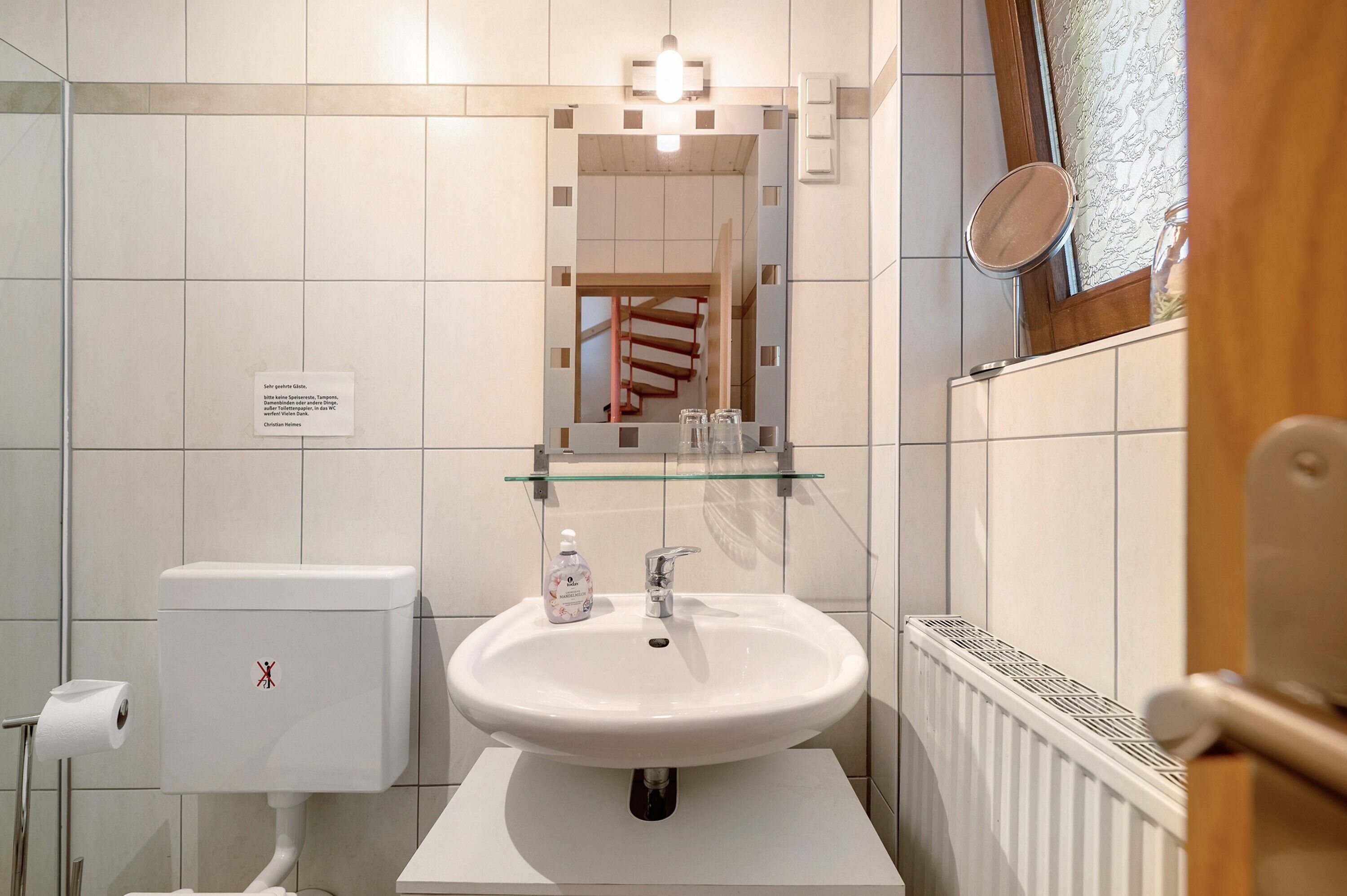 Une salle de bain avec un lavabo blanc, une étagère en verre au-dessus, des toilettes dotées d’un porte-serviettes en papier et un miroir.