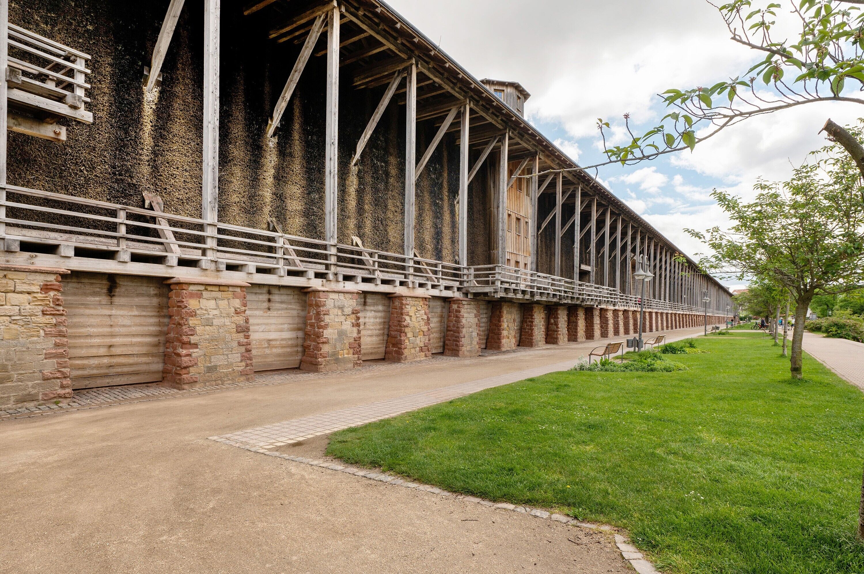 A long building with a brick facade and metal beams, a paved walkway, and a grassy area with a tree.