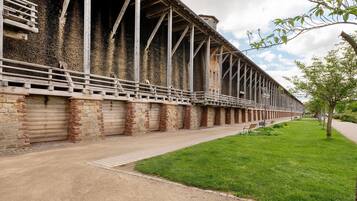 A long building with a brick facade and metal beams, a paved walkway, and a grassy area with a tree.