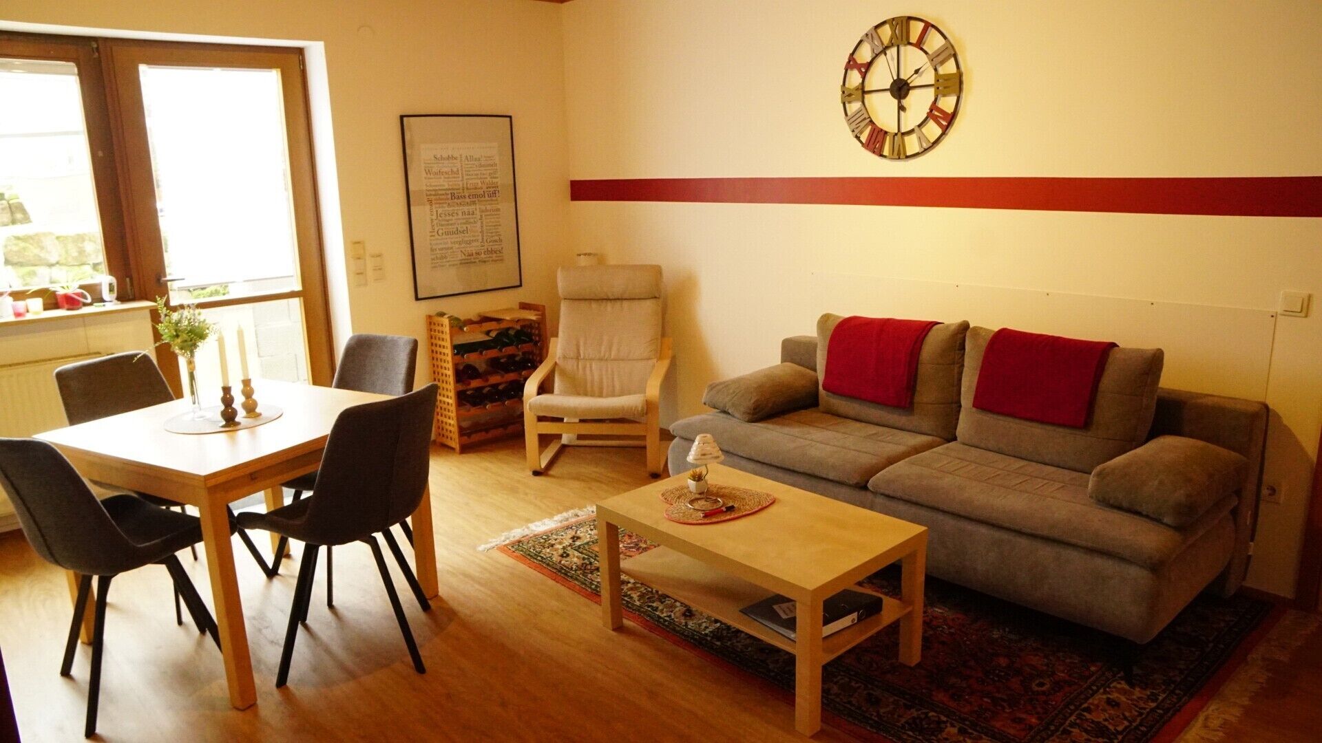Una sala de estar con sofá, una mesa de comedor con sillas, un reloj de pared y un menú enmarcado.