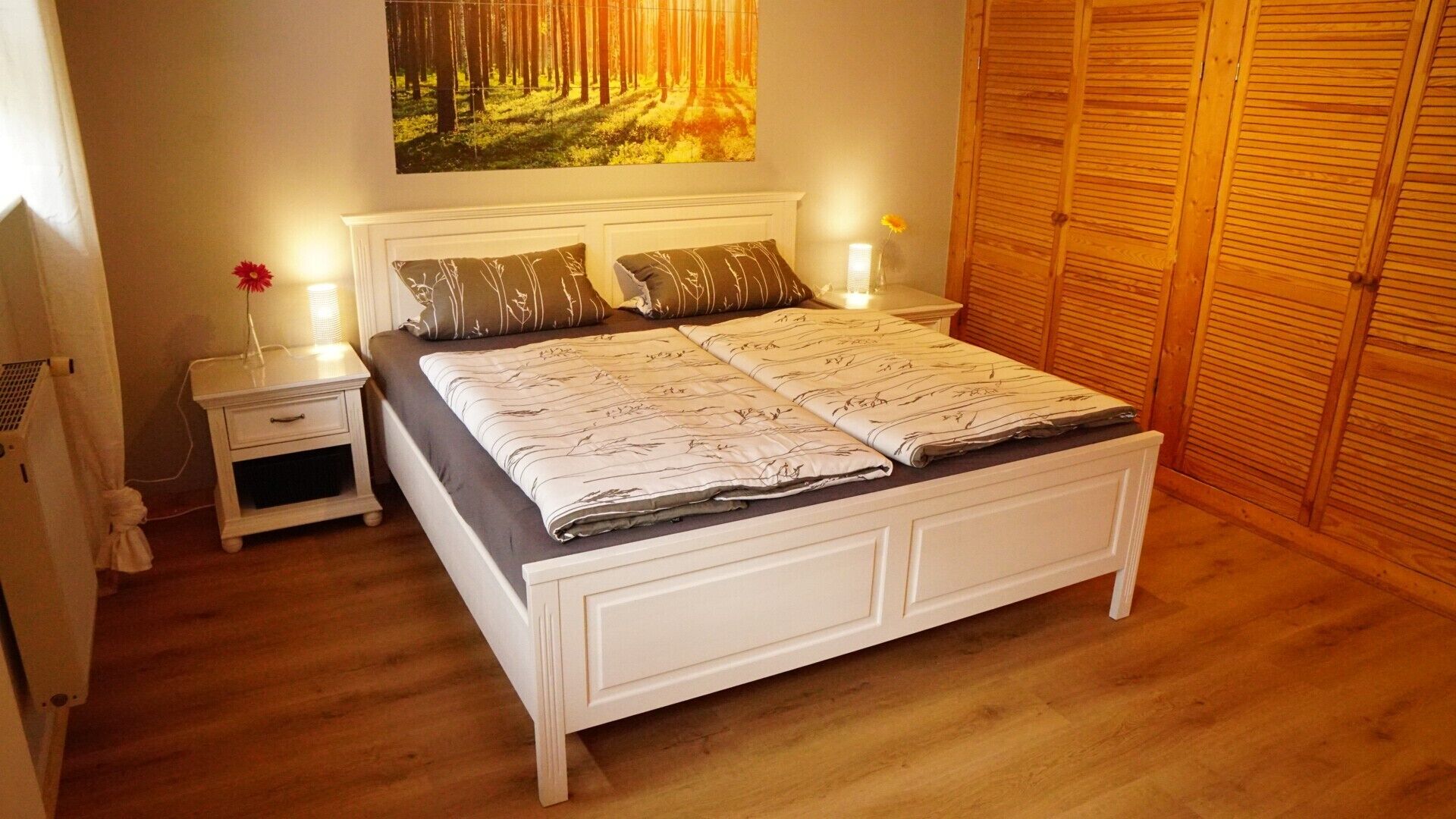 A bedroom with a bed, bedside tables, a nightstand, a wall-mounted painting, and a wooden wardrobe.