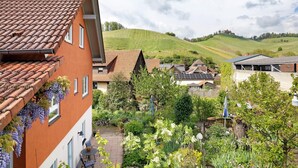 A residential area with terracotta-tiled roofs, a garden with various plants, and a hillside in the background.