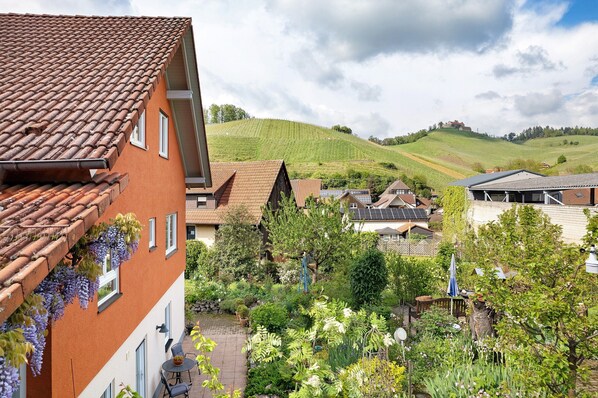 Ein Wohngebiet mit terrakottafarbenen Dächern, einem Garten mit verschiedenen Pflanzen und einer Hügelkulisse im Hintergrund.