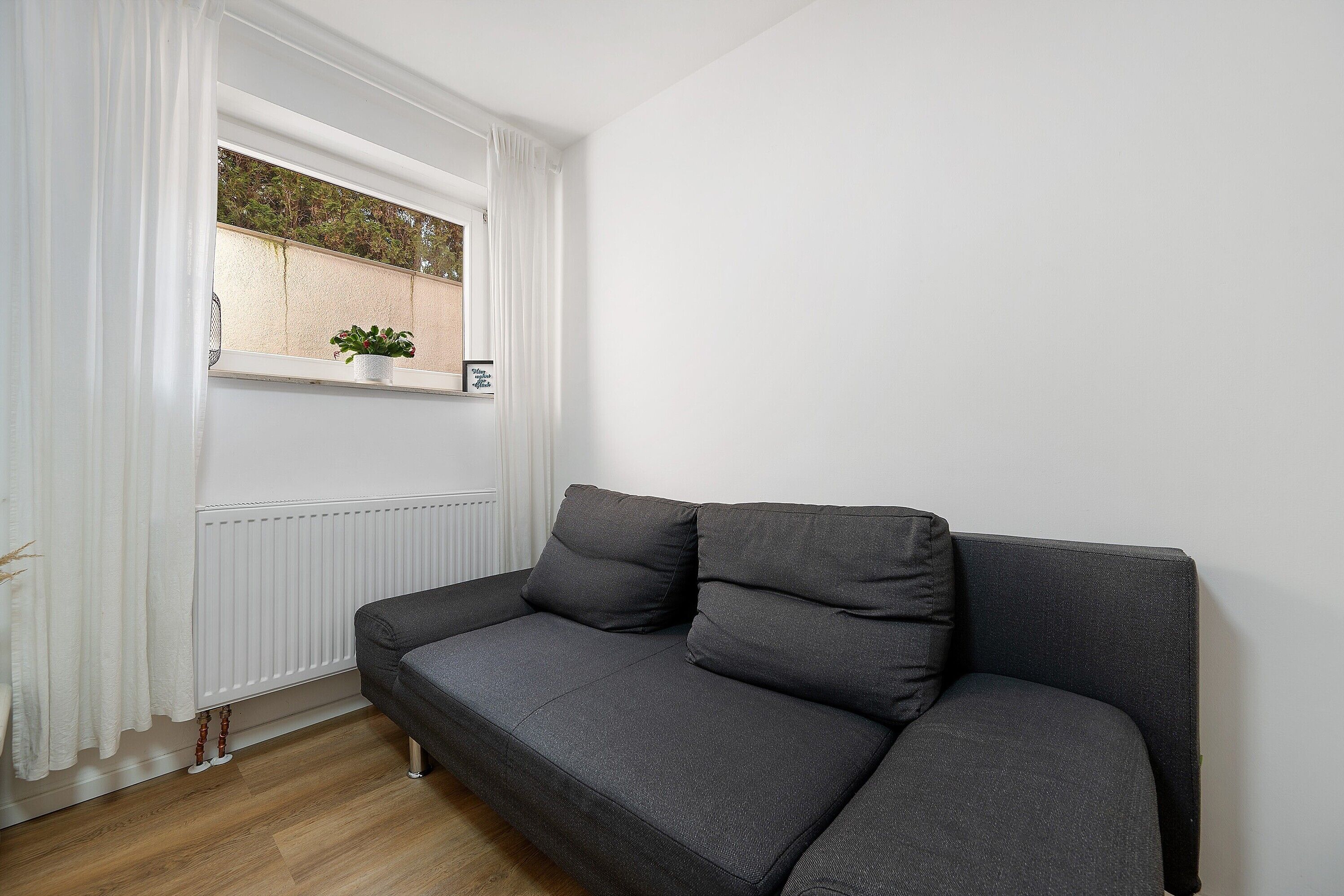 Un sofá gris con cojines, un radiador blanco, una ventana con una planta y una cortina blanca.
