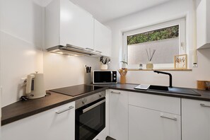 Una cucina moderna con mobili bianchi, un piano di lavoro nero, forno integrato, microonde e una finestra con vista su piante.