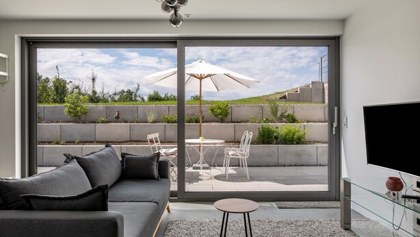 Una sala de estar moderna con un sofá, una mesa de centro de vidrio, un televisor y un ventanal corredero que da a un patio exterior con una mesa y sillas.