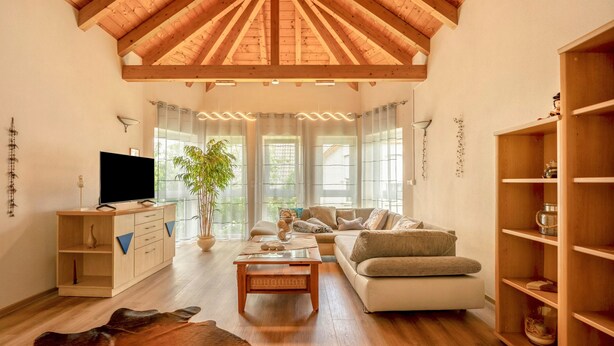 Ein geräumiges Wohnzimmer mit hoher Holzbalkendecke, einem Fernseher, einem Sofa, einem Couchtisch und einem Bücherregal.