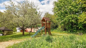 Eine hölzerne Spielkonstruktion mit Rutsche in einer grünen Rasenfläche, im Hintergrund Bäume und ein Schuppen.