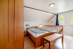 A bedroom with a wooden bed, a bedside table, a bench, a lamp, and a window with curtains.