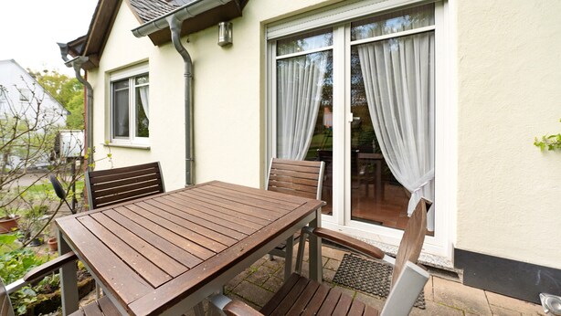 Eine Terrasse im Freien mit einem Holztisch und -stühlen, einer Glasschiebetür und einer weißen Außenwand.
