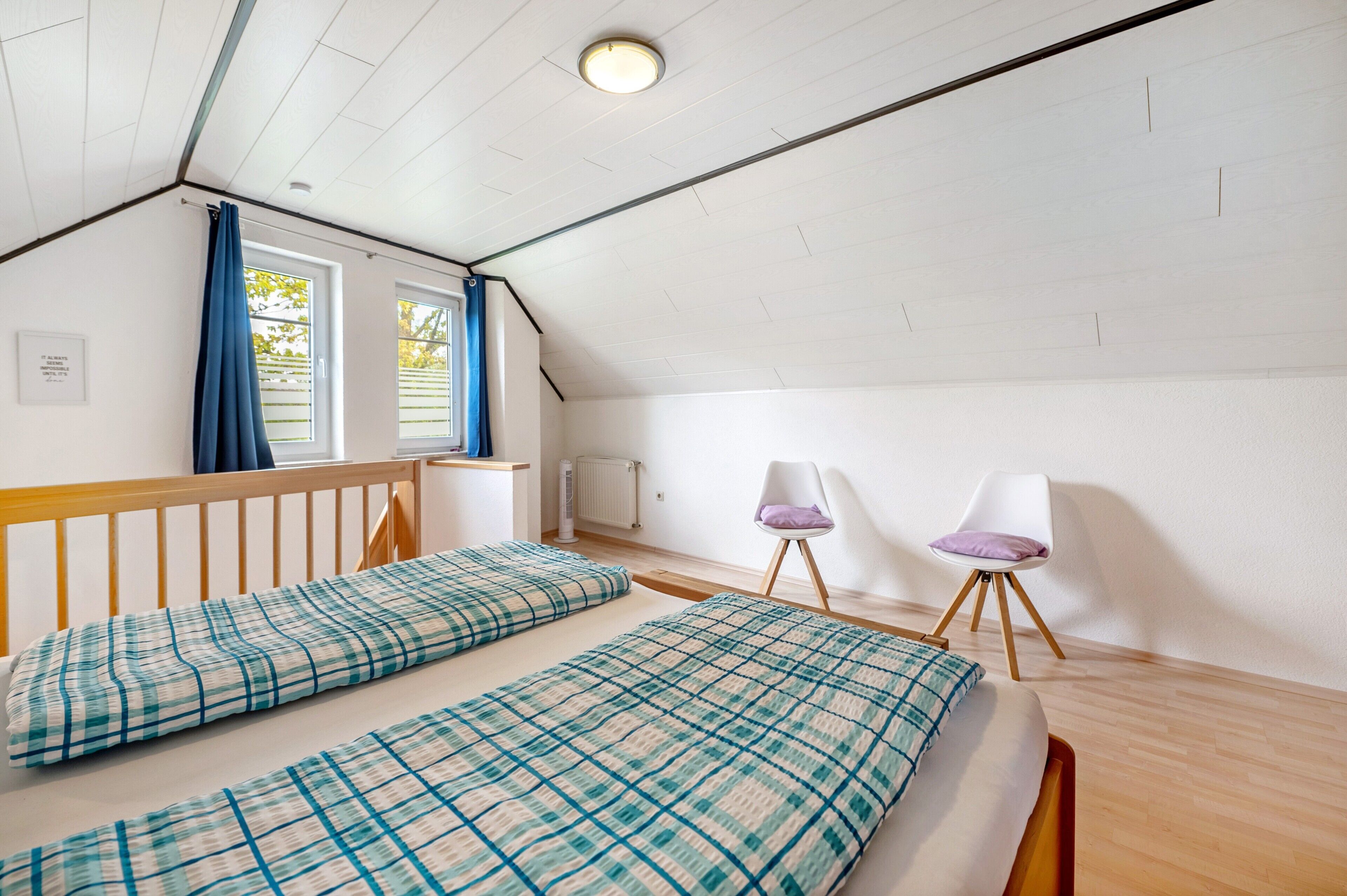 Ein Schlafzimmer mit schrägen Wänden, zwei Betten mit Karo-Bettwäsche, einem weißen Sessel und einem Fenster mit blauen Vorhängen.