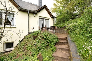 A house with a staircase leading to a door, surrounded by greenery and a brick wall.