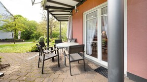 En udendørs terrasse med et bord og stole, et overdækket område og udsigt til en bygning med en glasdør.