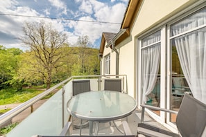 A balcony with a glass table and chairs, overlooking a garden and a building with large windows and curtains.