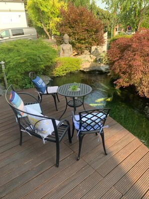 Um deck com duas cadeiras e uma mesa, um lago e uma estátua de Buda ao fundo.