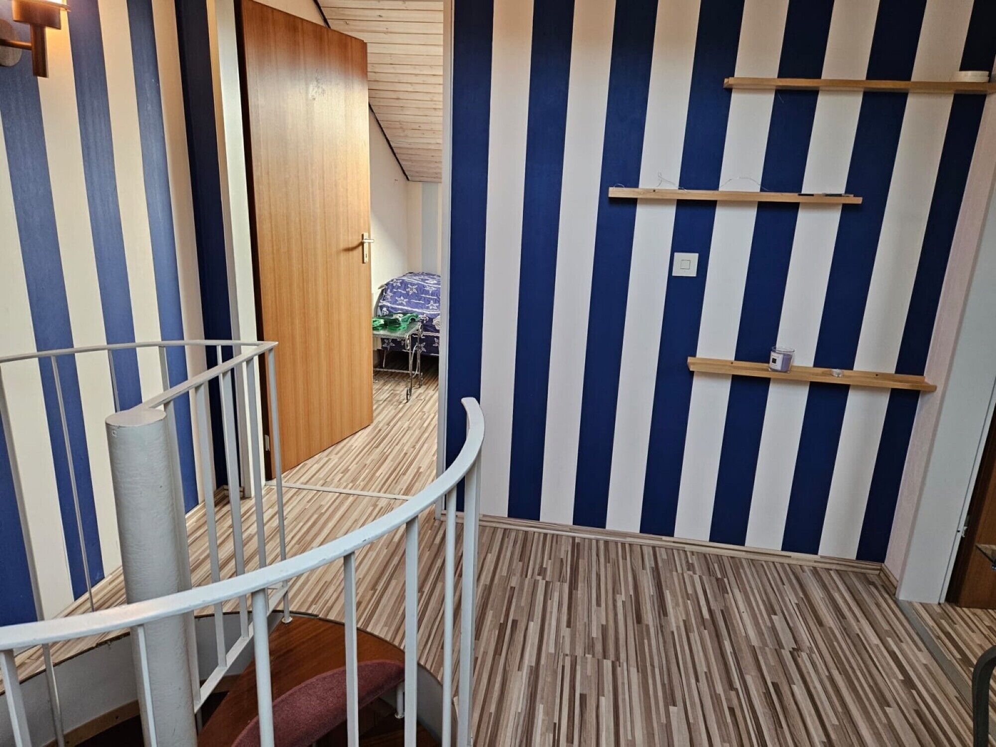 A room with a striped wall, a wooden door, a small table with a chair, and a white railing.