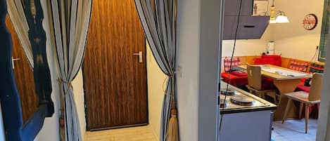 A room with a wooden door, a dining area with a red table, and a small kitchenette.