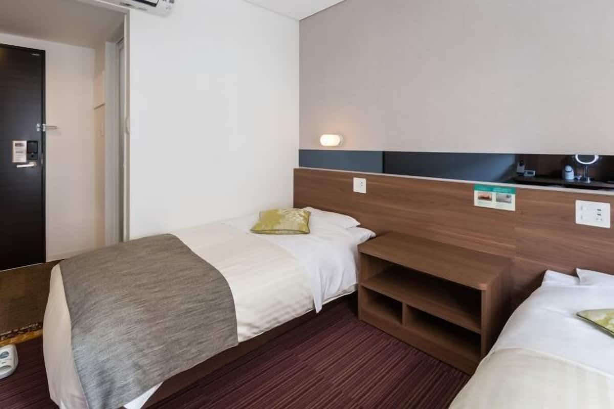 Une chambre d’hôtel avec un lit simple, une table de chevet, une petite étagère et une lampe fixée au mur.