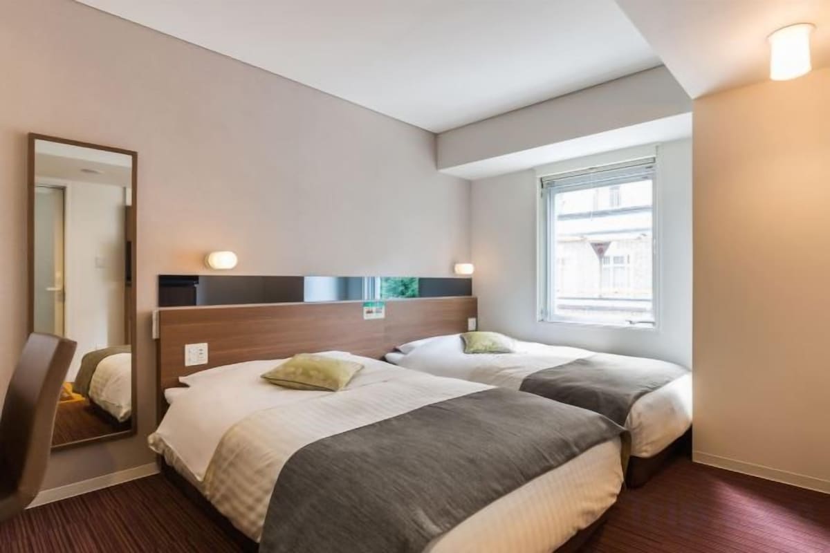 A hotel room with two beds, a mirror, a window with blinds, and a wall-mounted light fixture.