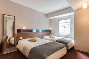 A hotel room with two beds, a mirror, a window with blinds, and a wall-mounted light fixture.
