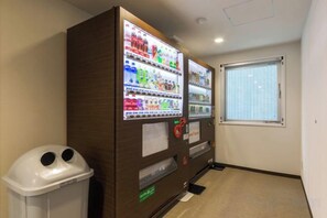 Un distributeur automatique proposant diverses boissons, une poubelle et une fenêtre avec des stores.