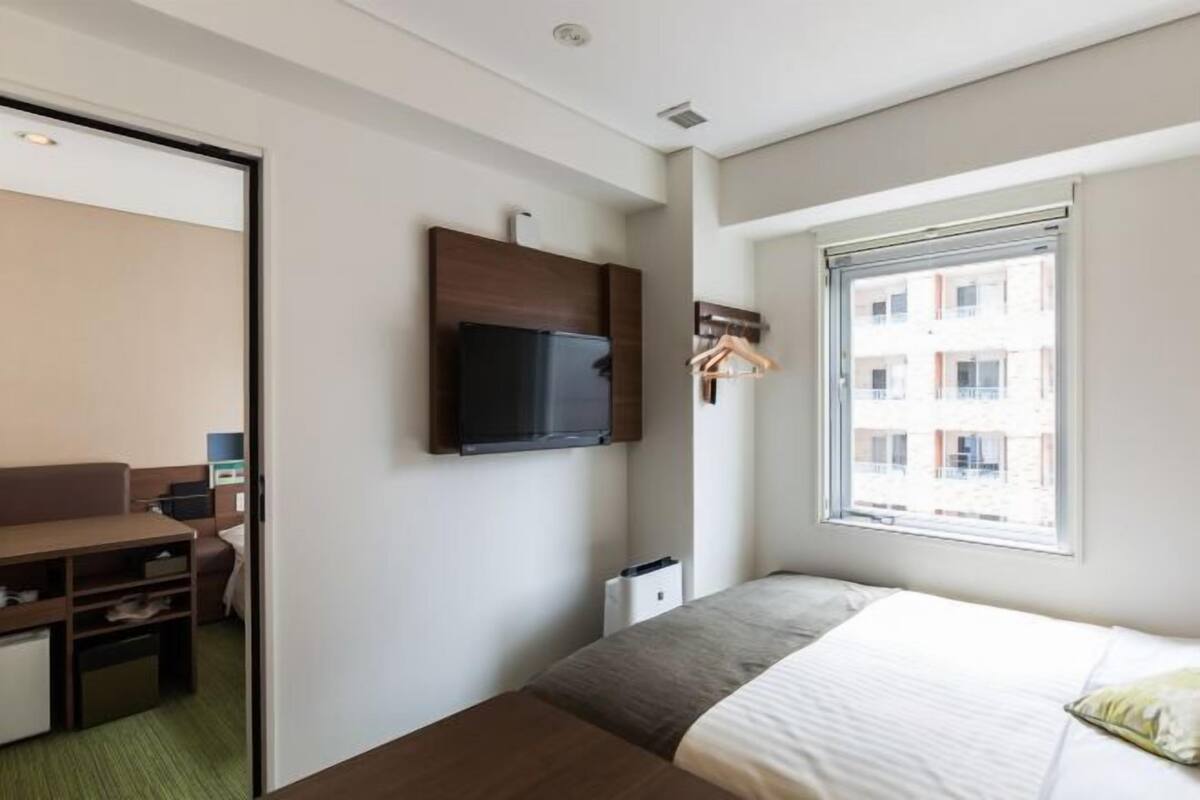 Une chambre d’hôtel avec un lit, un téléviseur fixé au mur, une fenêtre donnant sur des immeubles et un bureau avec une chaise.