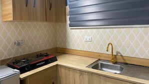 A kitchen with wooden cabinets, a gas stove, a sink with a gold faucet, and a window with horizontal blinds. - Fully Furnished Studio Apartment (Tema West Municipal)