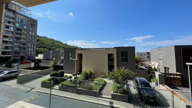 A modern urban residential area with a parking lot, a small building with a flat roof, and a multi-story apartment building in the background.