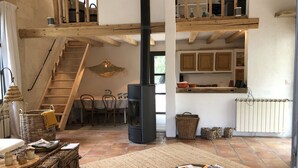 A spacious loft with a high ceiling, exposed wooden beams, a central fireplace, a dining area with a table and chairs, and a kitchen with wooden cabinets.