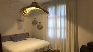 A bedroom with a bed, a hanging light fixture, a window with curtains, and a small wooden nightstand.
