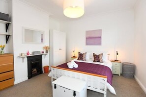 Quarto com cama, mesas de cabeceira, cômoda, espelho, lareira e um quadro na parede.