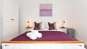 Un lit bien fait, avec une couvre-lit violette et deux coussins décoratifs, une porte blanche et une œuvre d’art accrochée au mur.