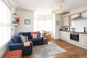 A modern living room with a blue sofa, a dining area with wooden chairs, and a kitchen with white cabinets and a microwave. - Modern Southville Apartment, 5 Min Walk to Centre (Bristol City)