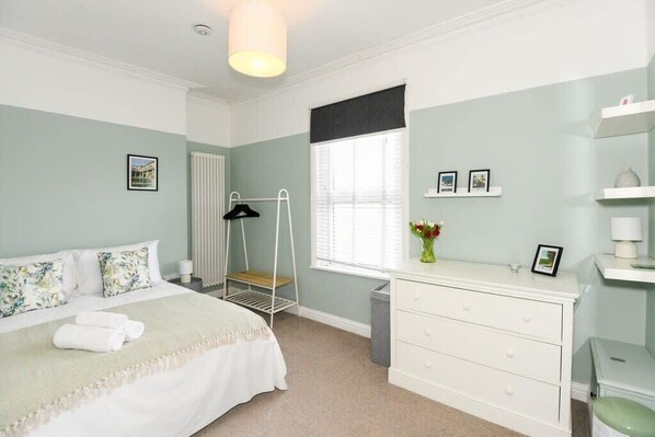 A bedroom with a bed, a bedside table, a radiator, a window with blinds, a hanging light fixture, and a small shelf with pictures.