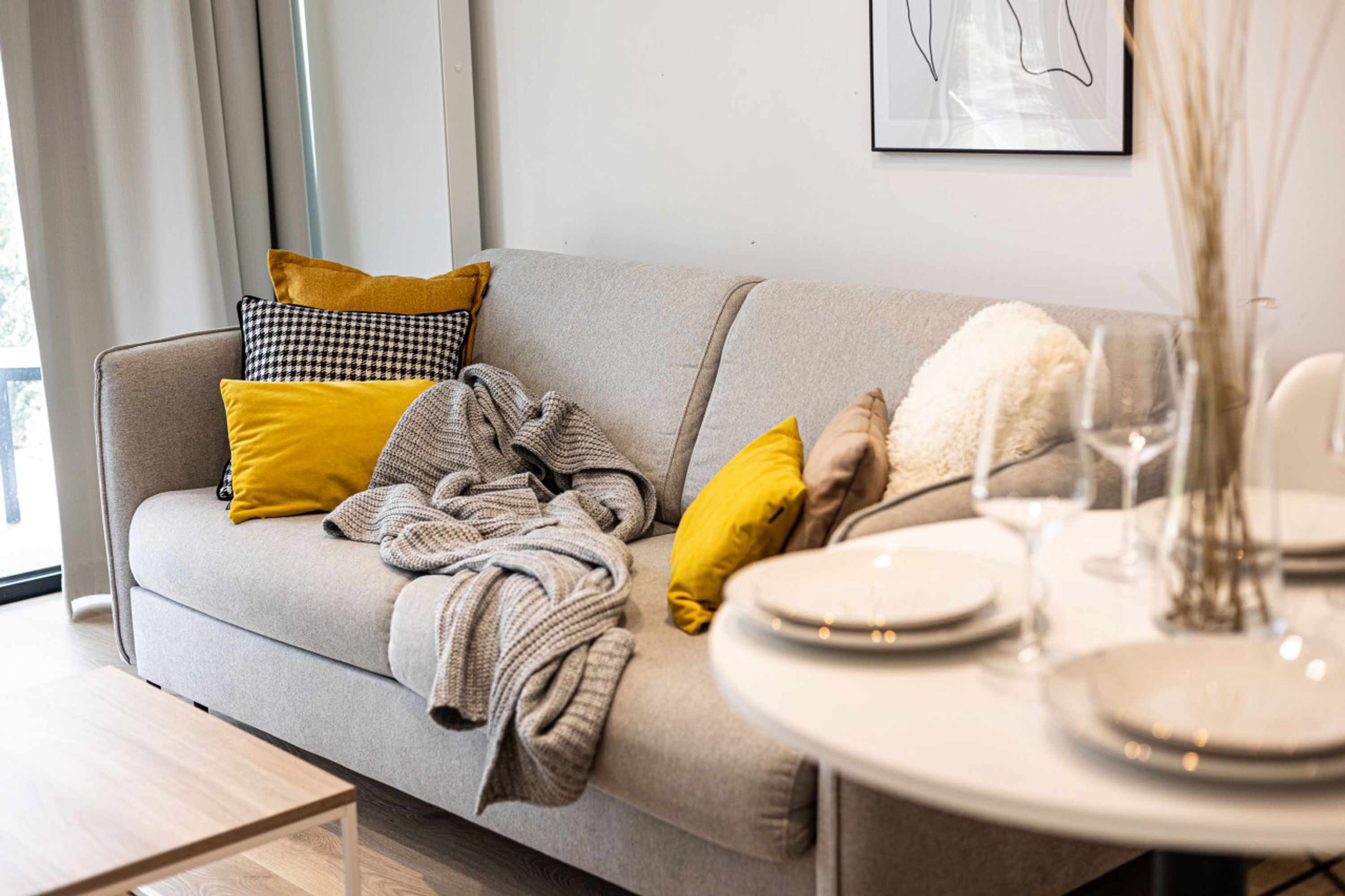 A modern living room with a grey sofa, yellow and black patterned pillows, a grey blanket, and a dining table set with plates and glasses.