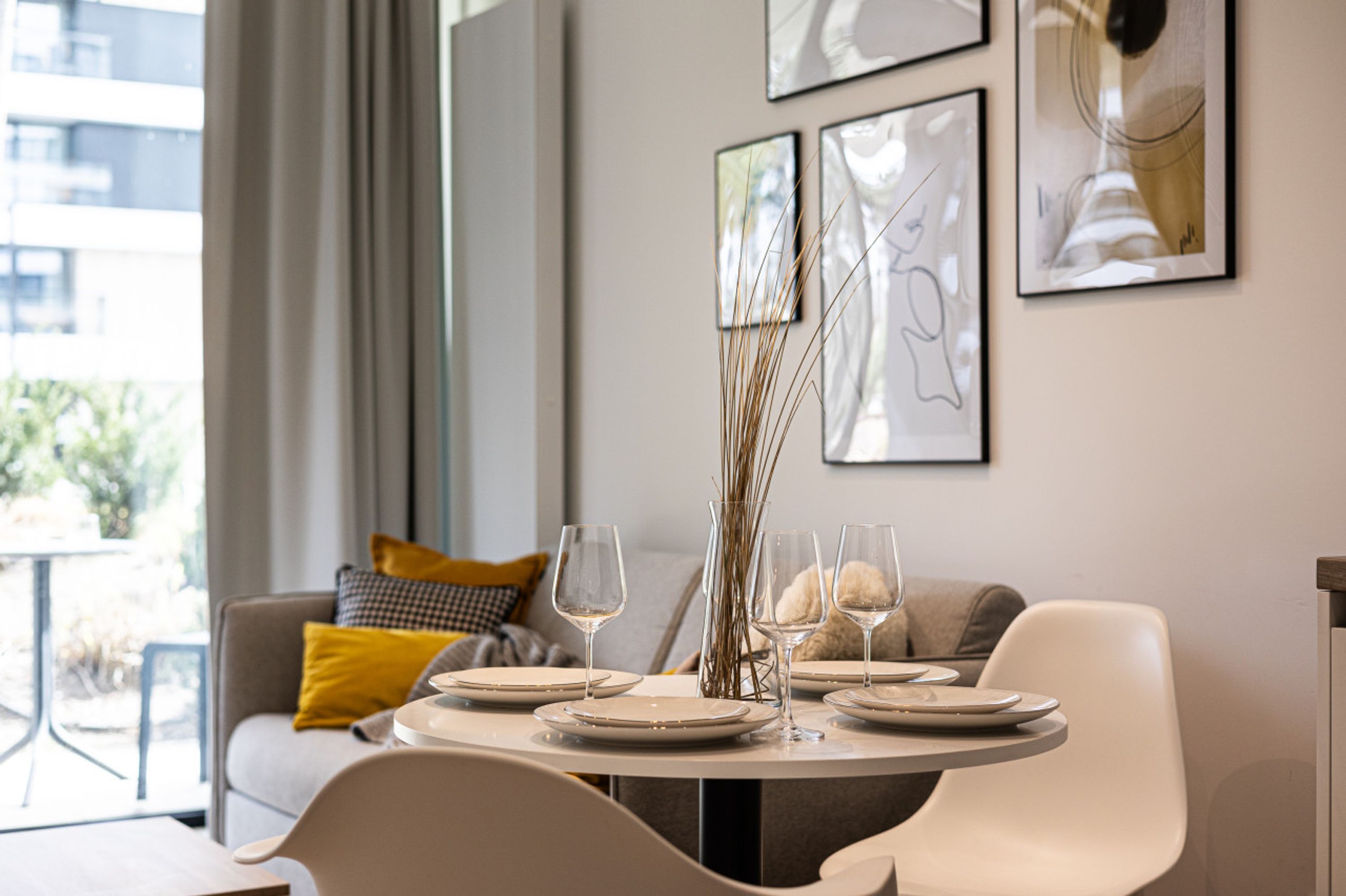 Un coin repas avec une table ronde dressée pour quatre personnes, agrémentée d’assiettes, de verres et d’un vase contenant de longues tiges de fleurs. La pièce présente une esthétique moderne, avec un canapé gris et un coussin jaune en arrière-plan.