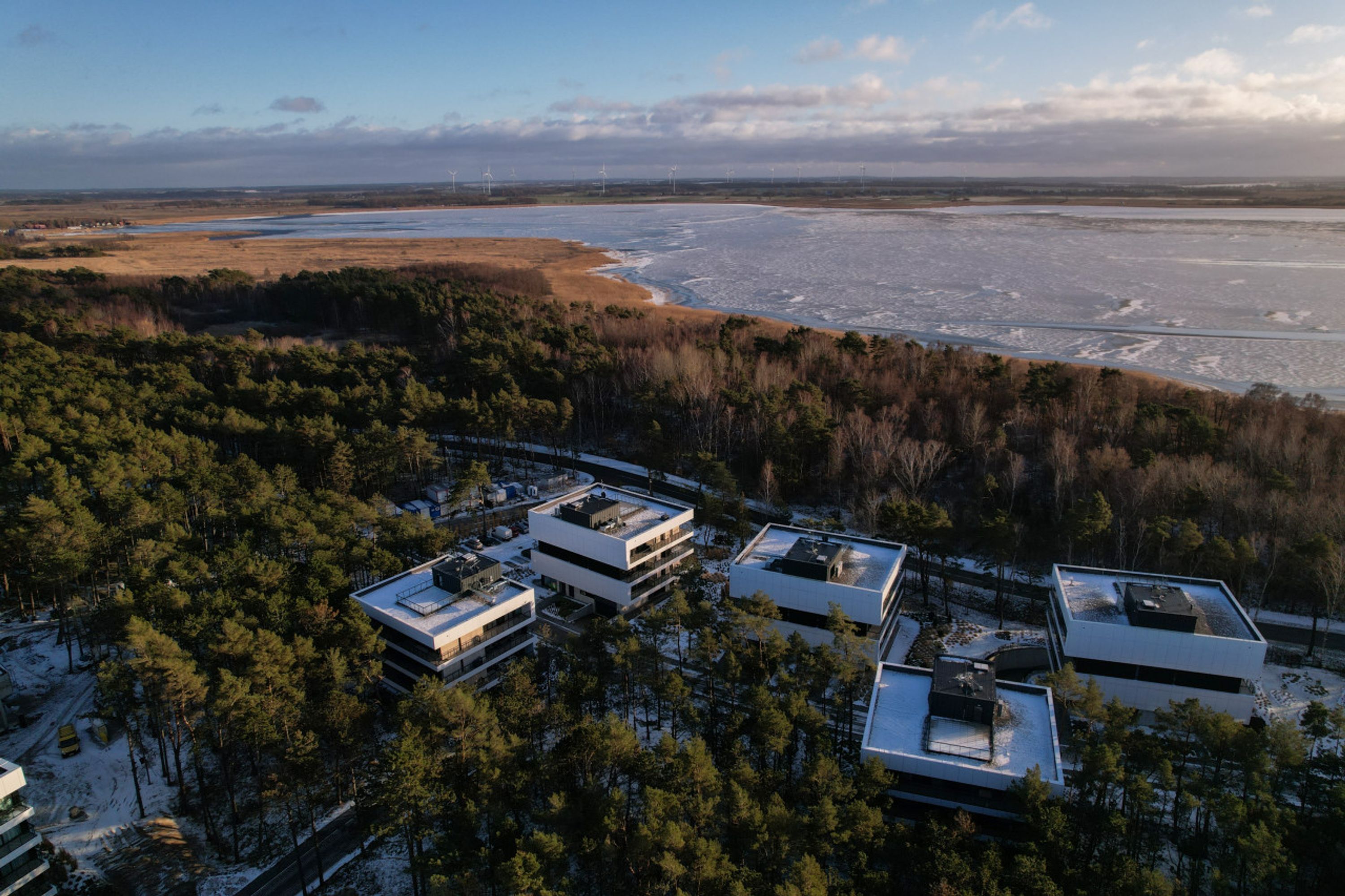 A modern building complex with multiple white structures, surrounded by dense forest and a frozen lake in the background.
