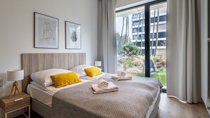 Une chambre d’hôtel moderne avec un grand lit, deux tables de chevet, une petite table et une fenêtre donnant sur un immeuble et de la verdure.
